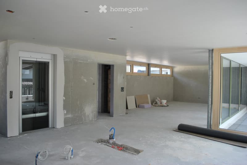 Empty room with unfinished concrete walls and floor, large glass doors, and scattered construction materials.