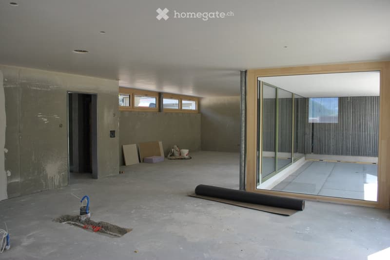 Empty room with unfinished concrete floor and walls, large glass window, and scattered construction materials including a rolled-up mat, buckets, and boards.