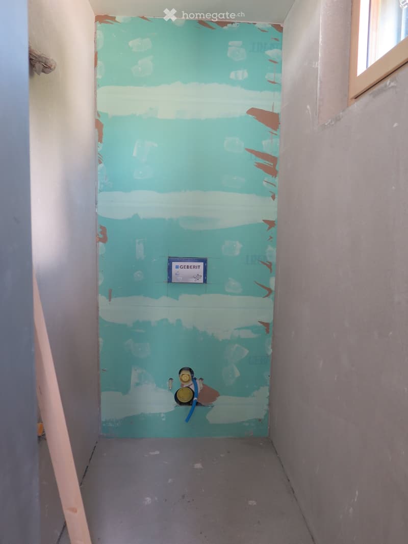 Empty room with unfinished walls, showing blue and white paint with exposed patches, a small window, and a water valve fixture on the floor.