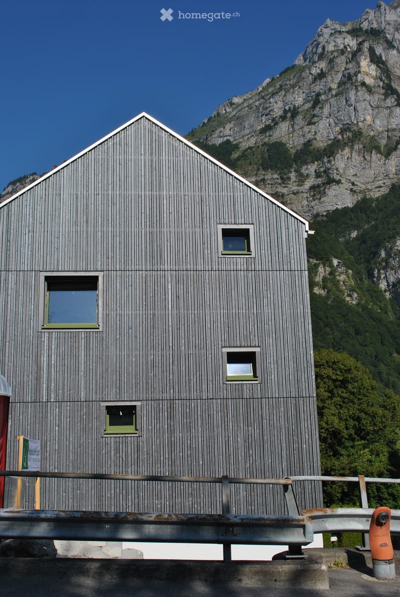 A modern, grey wooden building with a triangular roof and multiple windows, situated in a mountainous area with a clear blue sky and rocky terrain in the background.
