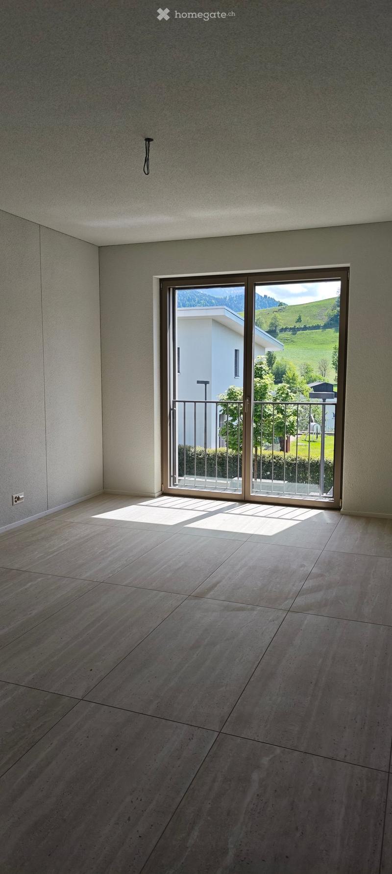 Empty room with light-colored tiled floor and large sliding glass door leading to a balcony with a view of green hills and a neighboring house.