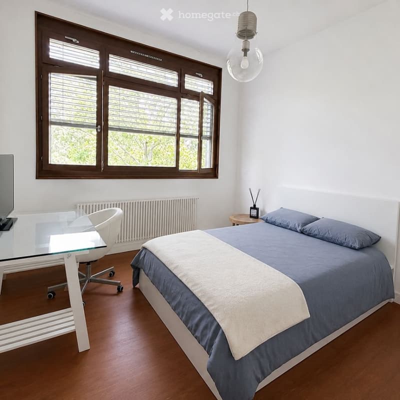 Modern bedroom with a large window, a bed with blue bedding, a glass desk, a white chair, and a small nightstand with a lamp.