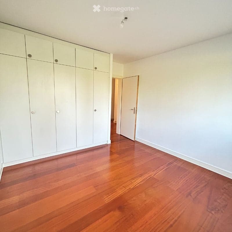 Empty room with light-colored walls, polished wooden flooring, and built-in white closet units. A door leads to another space.