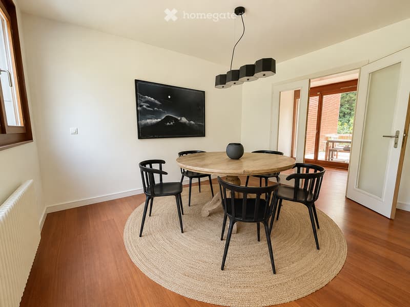 Dining area with a round wooden table surrounded by black chairs, a black vase as a centerpiece, a large picture frame on the wall, and a pendant light fixture above the table. The room has wooden flooring and a round woven rug under the table.