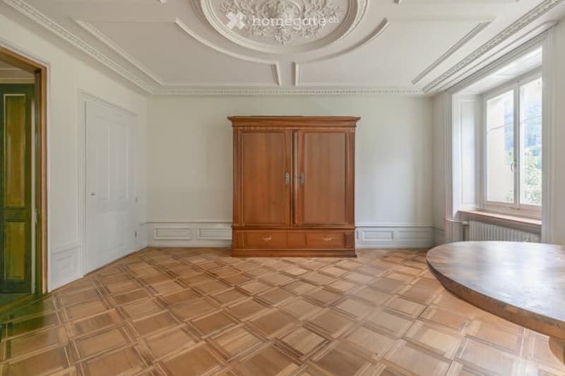 Elegant room with wooden parquet flooring, a large wooden wardrobe, and a round wooden table near the window.