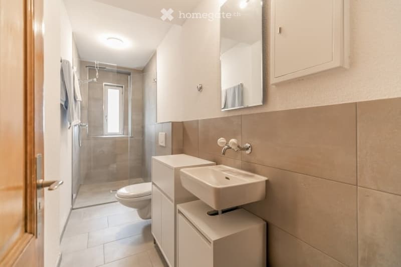 Modern bathroom with a white sink, toilet, mirror, and a glass-enclosed shower. Beige tiles and white cabinetry.