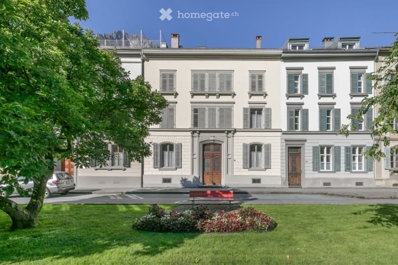 A multi-story residential building with classic architecture, featuring white walls, green shutters, and a central entrance. The building is surrounded by a well-maintained lawn with a red bench and flower beds.
