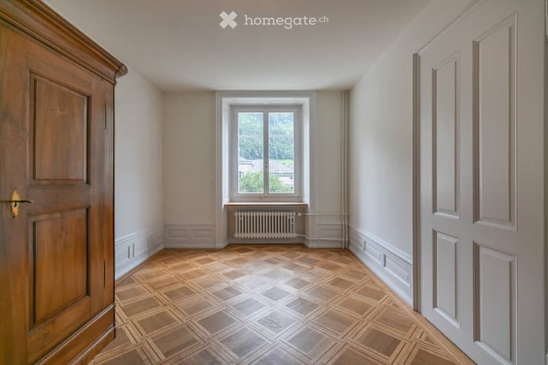 Empty room with wooden parquet flooring, white walls, and a window with a radiator below. There are two doors, one on each side of the room.
