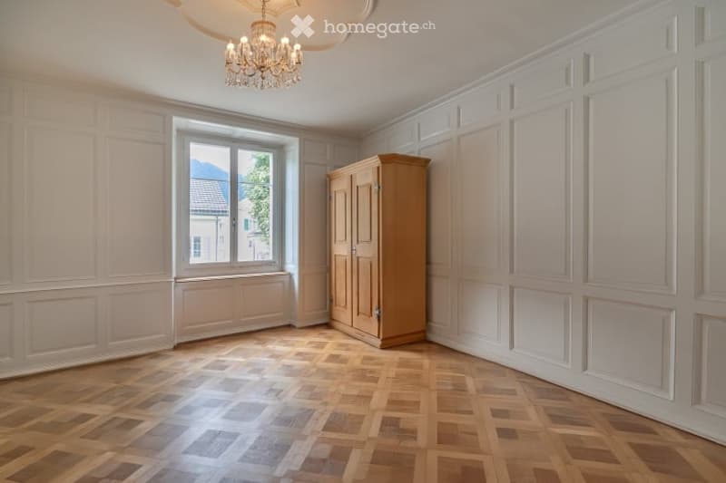 Empty room with wooden parquet flooring, white paneled walls, a large window with a view, and a simple wooden cabinet.