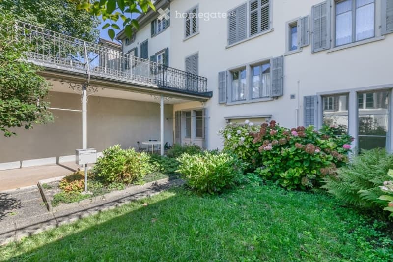 Well-maintained garden area with green grass, flowering shrubs, and a small patio with a table and chairs. A balcony with a metal railing is visible above the garden.