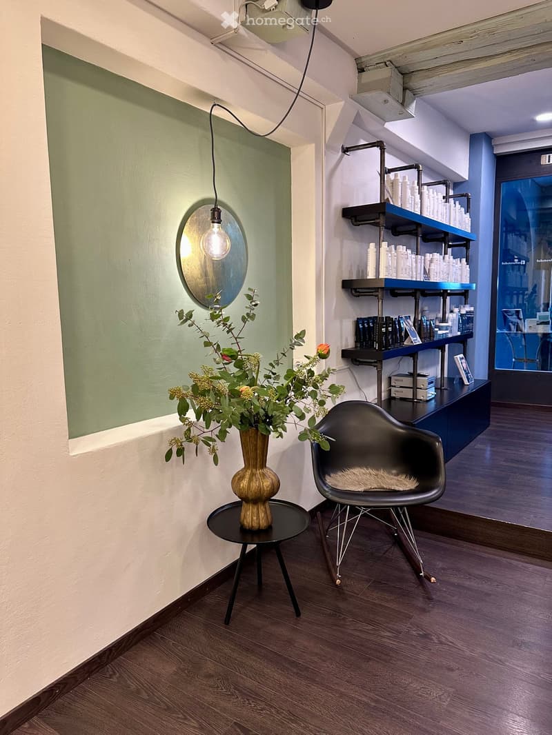 Modern interior with wooden flooring, a black chair with a fur cushion, a small round table with a golden vase holding a flower arrangement, shelves with various bottles, and a wall-mounted light fixture with a round mirror.