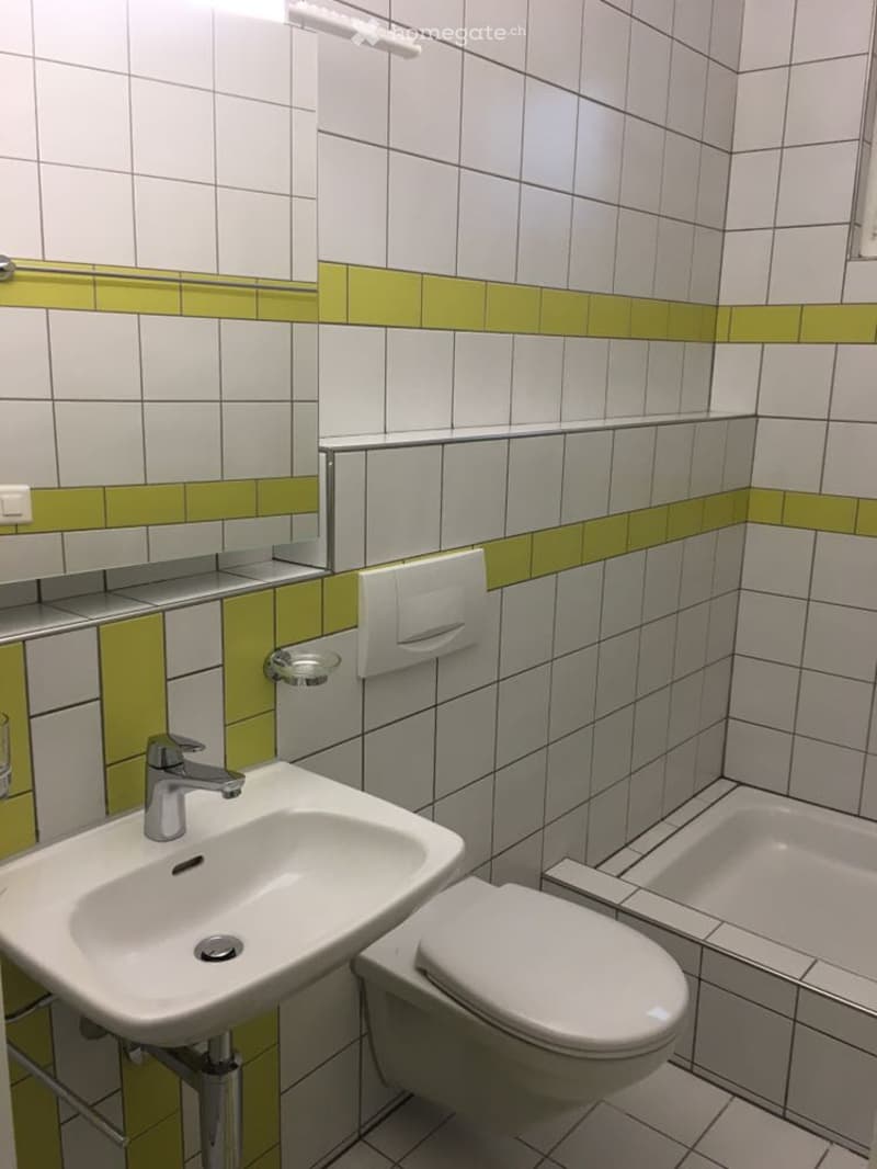 Modern bathroom with white tiled walls, green accent strip, sink, toilet, mirror, and towel rack.