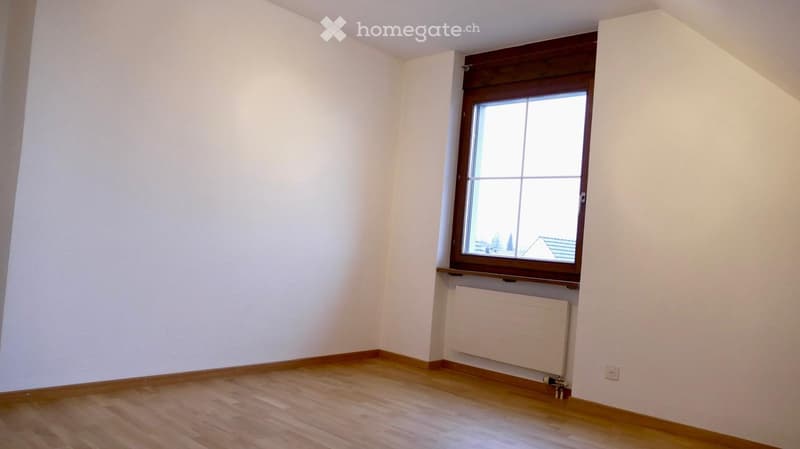 Empty room with wooden flooring, white walls, and a single window with wooden frame. Radiator is visible below the window.