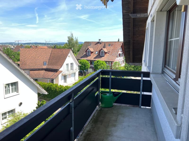 Balcony with a black railing, a green watering can, and a view of neighboring houses and greenery.