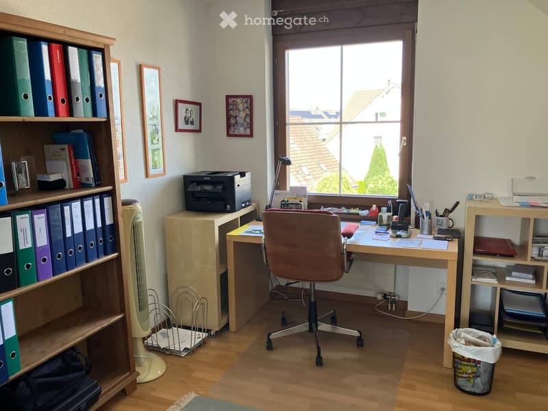 Office space with a wooden desk, office chair, printer, bookshelf, fan, and various office supplies.