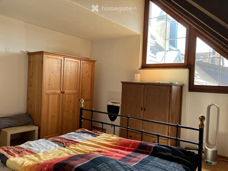 Cozy attic bedroom with wooden furniture, a bed with a colorful quilt, a wardrobe, and a nightstand with a fan.