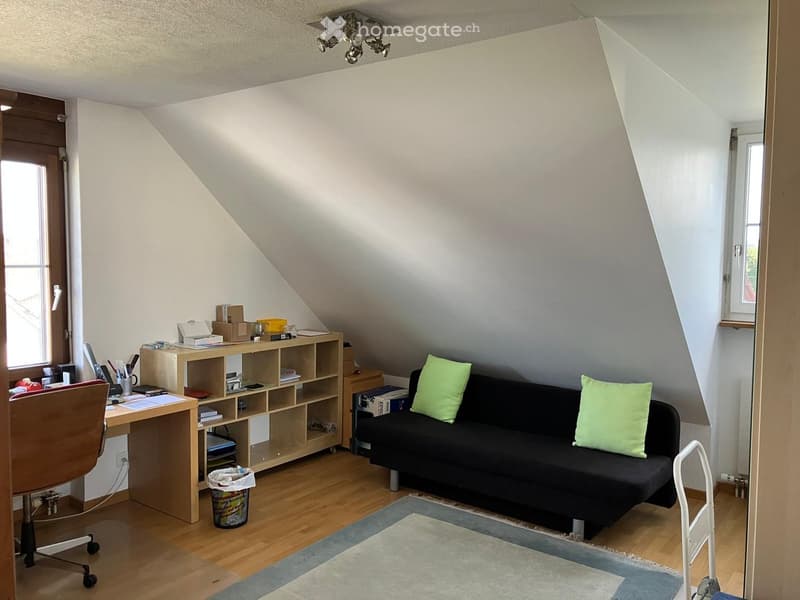 Cozy attic room with wooden flooring, slanted ceilings, and large windows. Features a black sofa with green cushions, a wooden desk with a chair, a shelf unit, and a small rug.