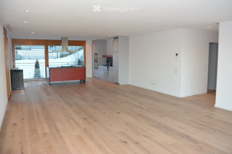 Open-plan kitchen and dining area with light wooden flooring, white walls, and modern cabinetry. Features include a large window with a view, a built-in kitchen island, and a stainless steel range hood.