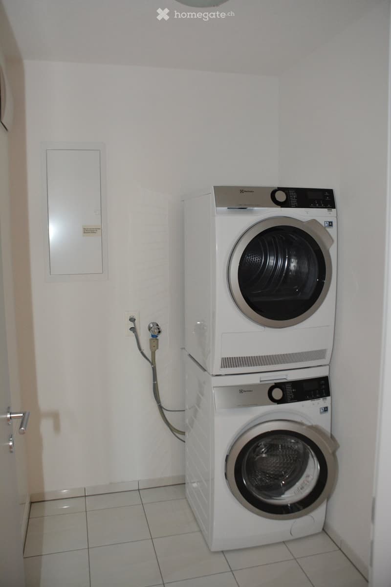 A small bathroom with a white tiled floor and white walls. A stacked washing machine and dryer unit is positioned against the wall, with a control panel and a door visible.