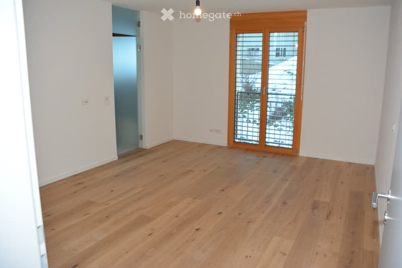 Empty room with light wooden flooring, white walls, and a large window with wooden frame and blinds. There is a visible electrical outlet and a door leading to another room.