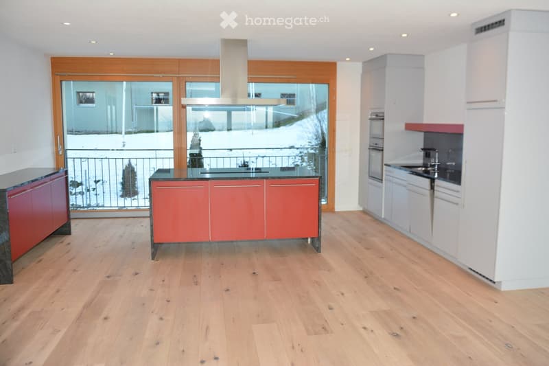 Modern kitchen with light wooden flooring, white and red cabinets, a central island, and large windows offering a view of the snowy outdoor area.