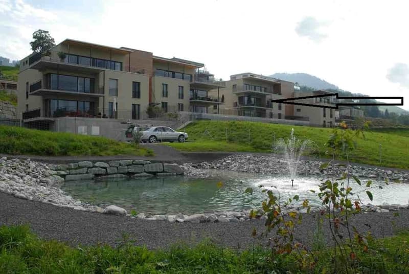 Modern multi-story residential building with balconies and large windows, situated on a hillside with a small pond and fountain in the foreground.