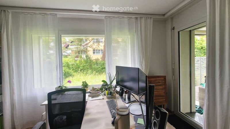 A well-lit home office with a desk, computer monitor, keyboard, chair, and several potted plants. Large windows with sheer curtains offer a view of greenery outside.