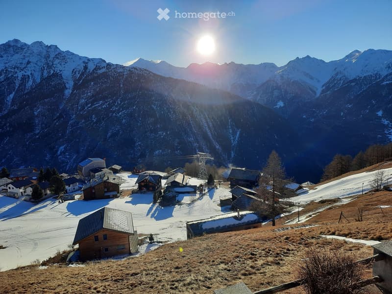 A picturesque village nestled in a snowy mountain valley, with scattered houses and a ski lift structure visible against a backdrop of snow-capped peaks.