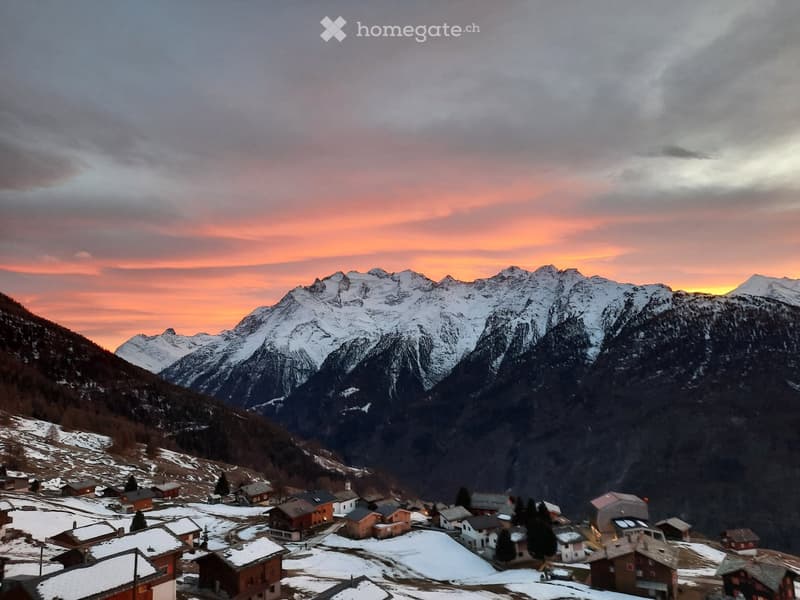 A picturesque mountain village at sunset, with snow-covered peaks and traditional wooden houses nestled in a valley.