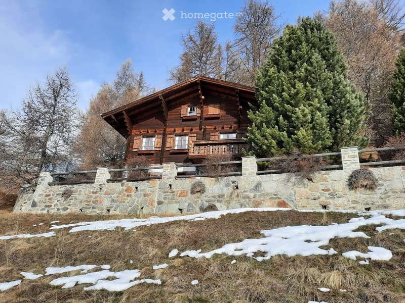 A two-story wooden house with a stone base, surrounded by trees and a stone wall, with patches of snow on the ground.