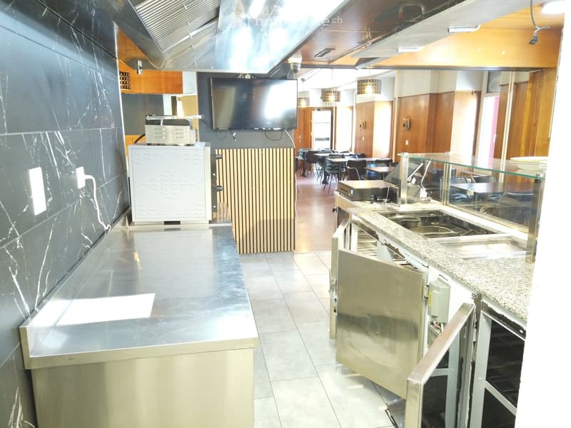 Commercial kitchen with stainless steel counters, refrigerator, oven, sink, and a television screen mounted on the wall.
