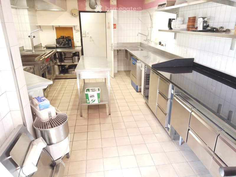 Commercial kitchen with stainless steel appliances, including a refrigerator, oven, sink, and mixer. Counters, shelves, and a small table are present.