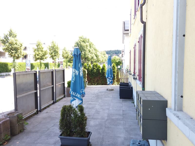 Balcony area with tiled floor, closed seating area with chairs and umbrellas, potted plants, and a view of greenery and buildings.