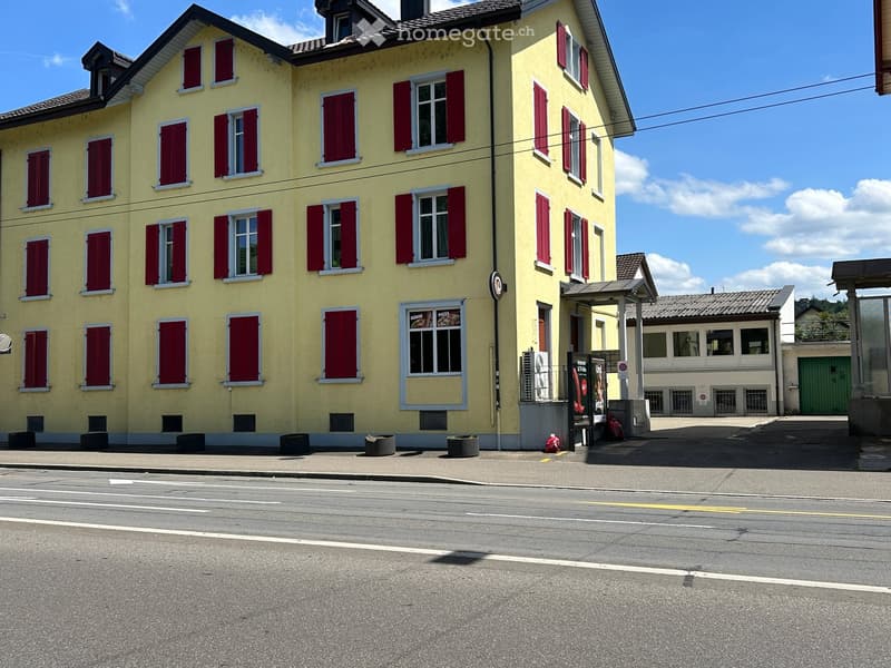 Two-story yellow building with red shutters and multiple windows, located on a street corner with a clear blue sky and some scattered clouds in the background.