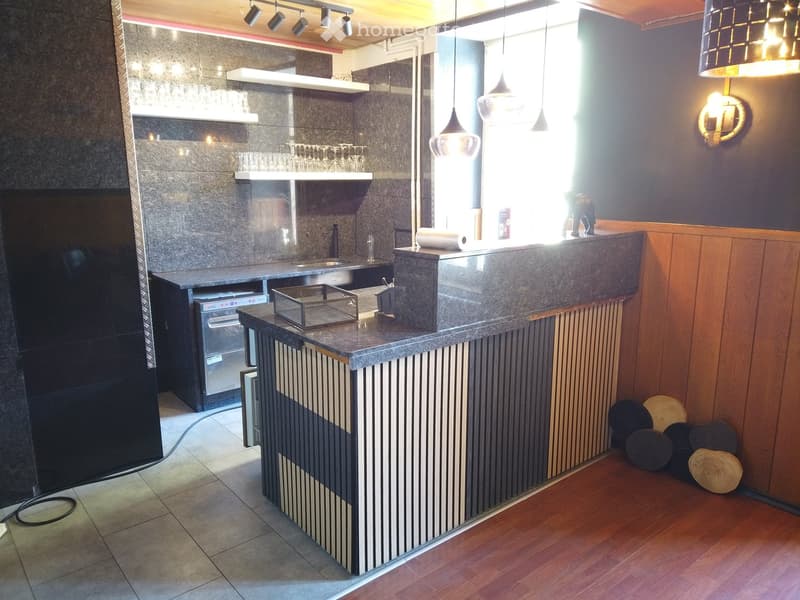 Modern reception area with a stone countertop, sink, refrigerator, shelves, pendant lights, and a small seating area with stools.