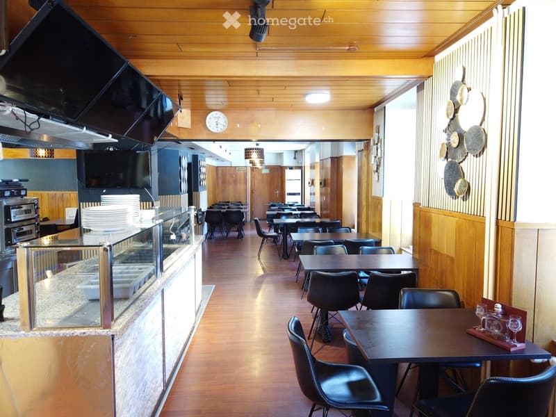 Restaurant interior with wooden paneling, tables, chairs, a counter with dishes, and a TV screen. The space is well-lit with natural light and features decorative wall plates.