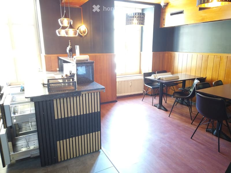 A modern bar area with a central counter, bar stools, and a dining area with tables and chairs. The space features wooden flooring, pendant lighting, and a refrigerator.