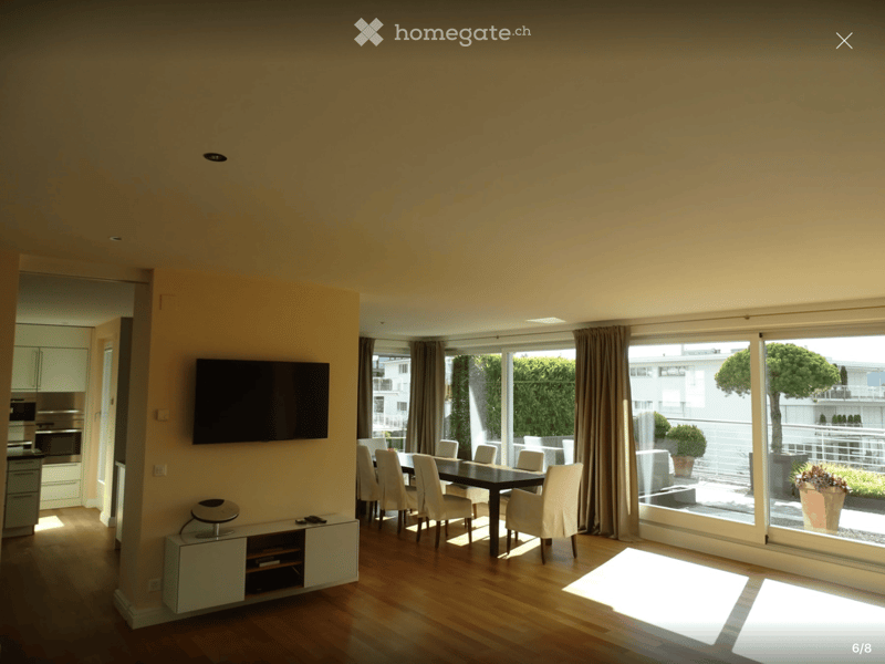 Bright living and dining area with wooden flooring, large windows, a flat-screen TV, a dining table with chairs, and a small cabinet with a stool.