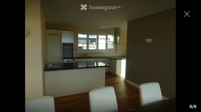 Modern kitchen with white cabinets, a central island, and a dining area with white chairs and a table. The kitchen features a sink, oven, and a bottle on the counter.