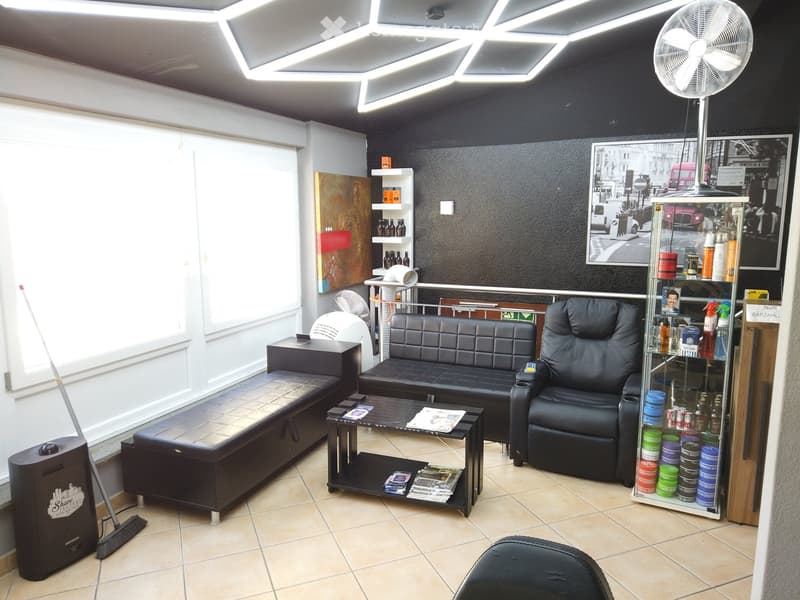 Modern interior with black leather sofa, armchair, coffee table, shelves with bottles, and a large fan. The room has large windows with blinds and a tiled floor.