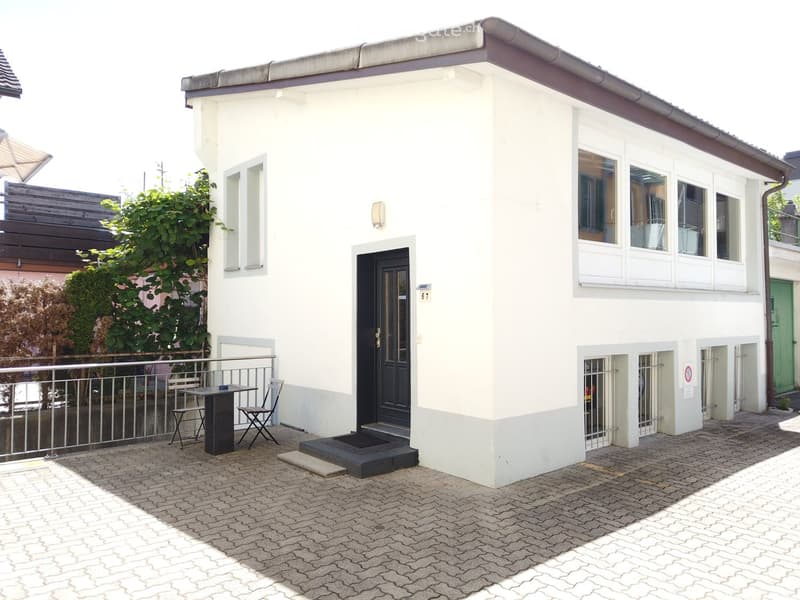 Two-story white building with a small outdoor seating area featuring a table and chairs, a metal railing, and a paved cobblestone area.