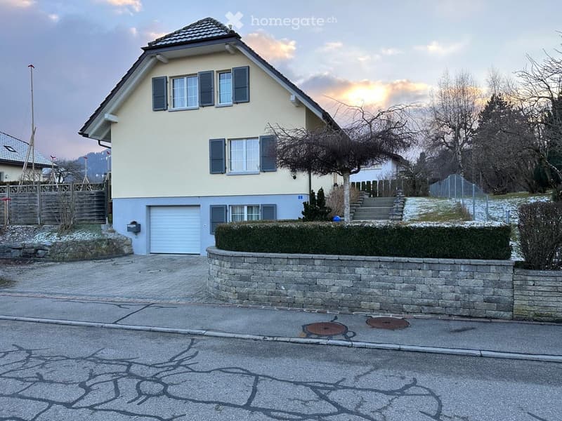 Two-story house with beige exterior, blue shutters, garage, and stone wall in front.