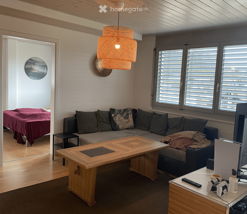 A living room with a sofa, coffee table, and a lamp. There is a bed in the adjacent room. A remote control, bottle, and game controllers are on the desk.