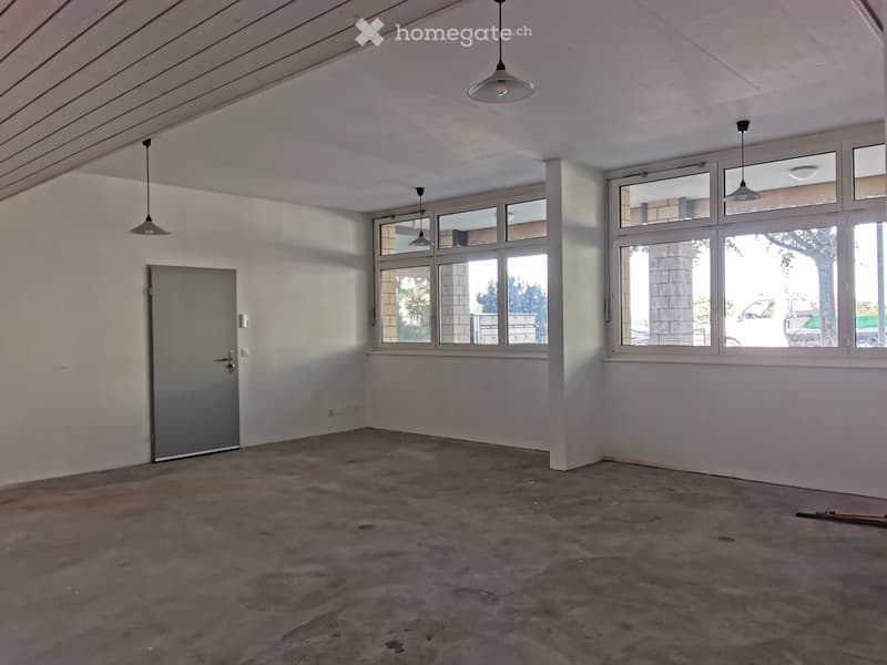 Empty room with concrete floor, white walls, large windows, and pendant lights.
