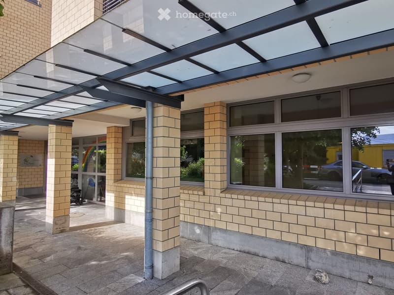 Modern building entrance with a glass canopy supported by brick pillars, large windows, and a paved ground floor area.