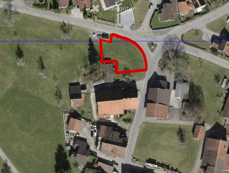 Aerial view of a residential area with multiple houses, roads, and green spaces. A red outline highlights a specific plot of land.