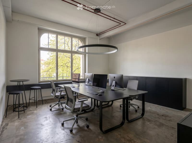 Modern office space with a large table, multiple chairs, and computer screens, illuminated by a circular pendant light and large windows.