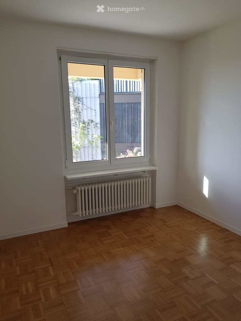 Unfurnished Room. Empty room with wooden flooring, white walls, large window with white frame, radiator below the window, and a small shelf above the radiator.