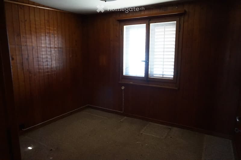 Empty room with wooden panel walls and a single window with blinds. The floor is carpeted, and there are no visible furnishings or decorations.