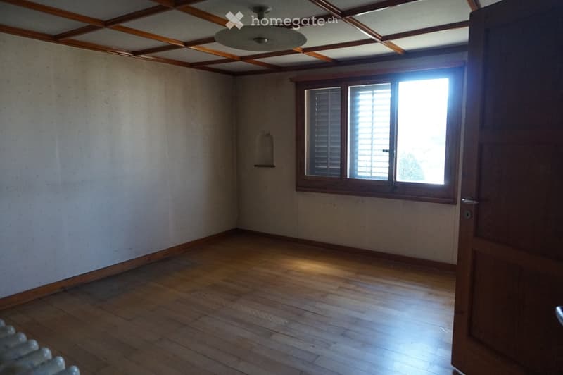Empty room with wooden flooring, white walls, and a large window with blinds. The ceiling features exposed wooden beams and a central light fixture.